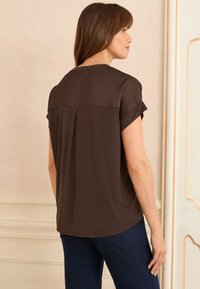 Hnědá blůza s krátkým rukávem a kulatým výstřihem, se záševkem na zadní straně a jemnou texturou. V kombinaci s tmavými džíny.