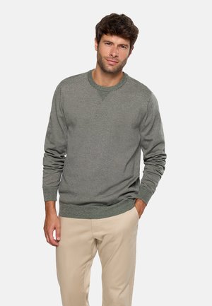 Hombre que lleva un suéter gris de cuello redondo y pantalones beige, de pie con una mano en el bolsillo contra un fondo blanco liso.