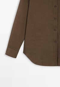 Chemise marron à manches longues avec boutons, une seule manche avec poignet et ourlet incurvé sur fond blanc.