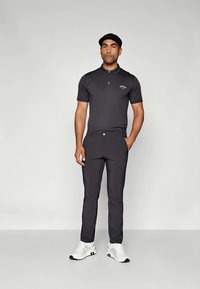 Mand iført sort polo skjorte, sorte bukser, hvide sneakers og sort flad cap står foran en grå baggrund.