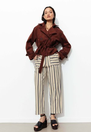 Femme debout contre un mur blanc, portant une veste marron ceinturée, un pantalon crème à jambes larges avec des rayures verticales noires et des sandales compensées noires.