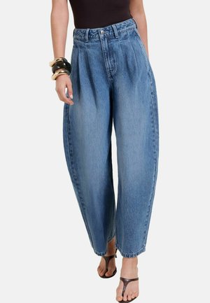 Frau trägt locker sitzende blaue Jeans mit hoher Taille, schwarzes ärmelloses Oberteil, schwarze und goldene Armbänder sowie dunkelbraune, offene Sandaletten mit Absatz.
