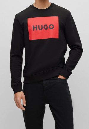 Man in een zwart sweatshirt met een rode vierkant en vetgedrukte "HUGO" tekst, gecombineerd met zwarte broek, staande tegen een grijze achtergrond.