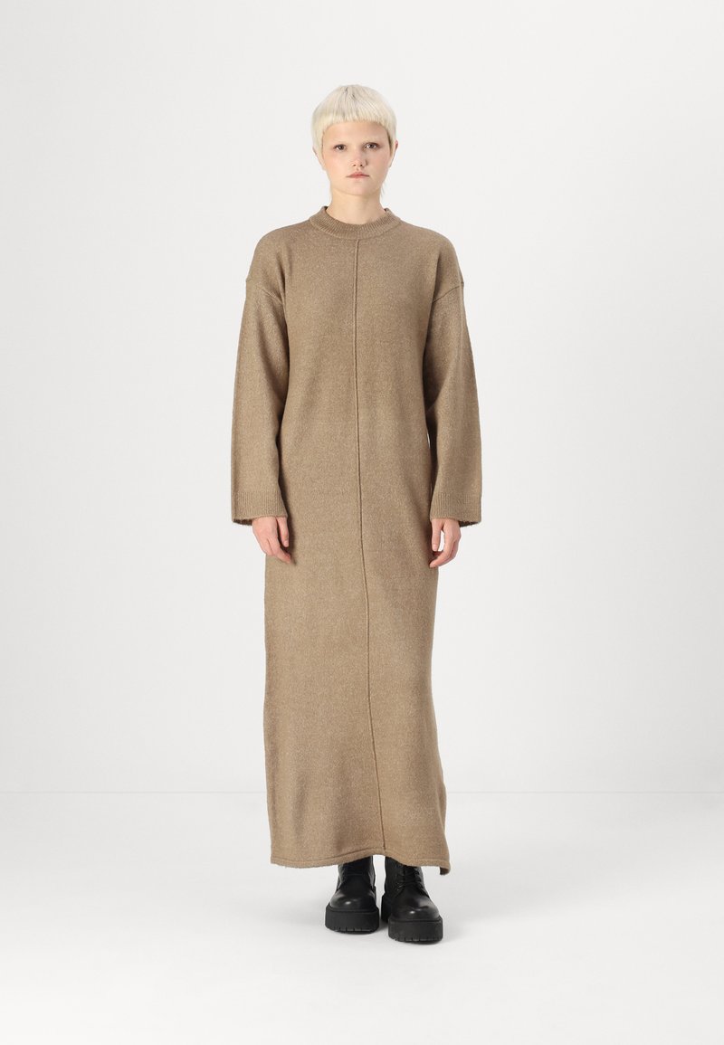 Robe en tricot beige à manches longues avec une coupe droite, en tissu texturé et un col montant. Portée avec des bottes à plateforme noires.