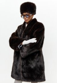 Faux fur frakke i mørkebrun med bred krave, lange ærmer og afslappet pasform. Sammen med en matchende pels hat og hvide læderhandsker.