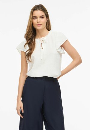 Kobieta z długimi włosami, ubrana w białą bluzkę z krótkim rękawem i wiązanym kołnierzem oraz granatowe, szerokie spodnie z wysokim stanem na gładkim tle.