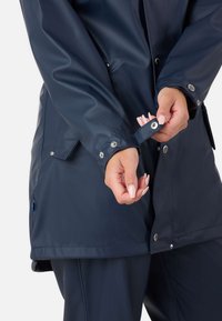 Personne fermant le bouton-pression du poignet d’une veste imperméable bleu foncé, montrant des détails métalliques brillants et une texture de tissu lisse.