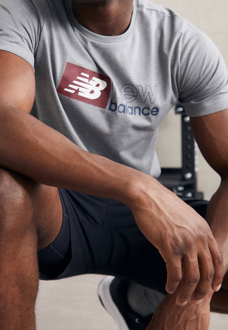 Graues Sport-T-Shirt mit großem roten und blauen "New Balance"-Logo, kurzen Ärmeln und eng anliegendem Schnitt. Texturiertes Material, kombiniert mit schwarzen Shorts.