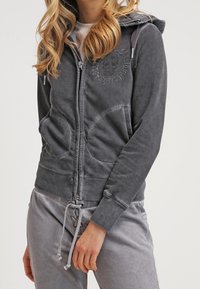 Sweat à capuche zippé gris avec une capuche, des poches avant et un graphique de crâne. Fabriqué dans un matériau doux avec une coupe décontractée et des détails à cordon.