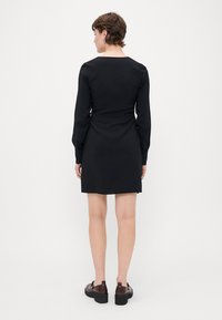 Melna garās piedurknes kleita ar piegulošu dizainu, apaļu kakla izgriezumu un pogu detaļām uz piedurknēm, apvienota ar tumši brūnām platformas kurpēm.