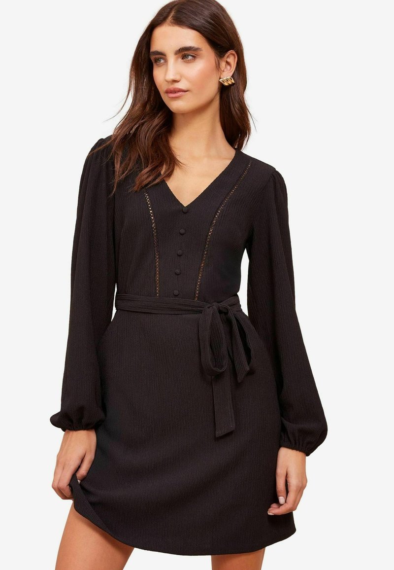 Schwarzes geripptes Kleid mit langen, gepufften Ärmeln, V-Ausschnitt, Frontknöpfen und einem Bindegürtel in der Taille. Strukturiertes Gewebe mit zarten Details.