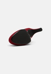 Chaussure à talon haut rouge montrée par la semelle, avec un dessous pointu noir et un talon fin sur fond blanc uni.