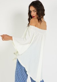 Blusa blanca sin mangas con escote recto, cuello smock texturizado, mangas acampanadas fluidas y dobladillo con encaje, combinada con pantalones azules con estampado.