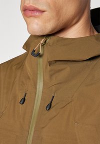 Veste brun olive avec un col haut, fermeture éclair à l'avant et cordons de serrage réglables. Présente des coutures détaillées et un tissu texturé pour plus de durabilité.