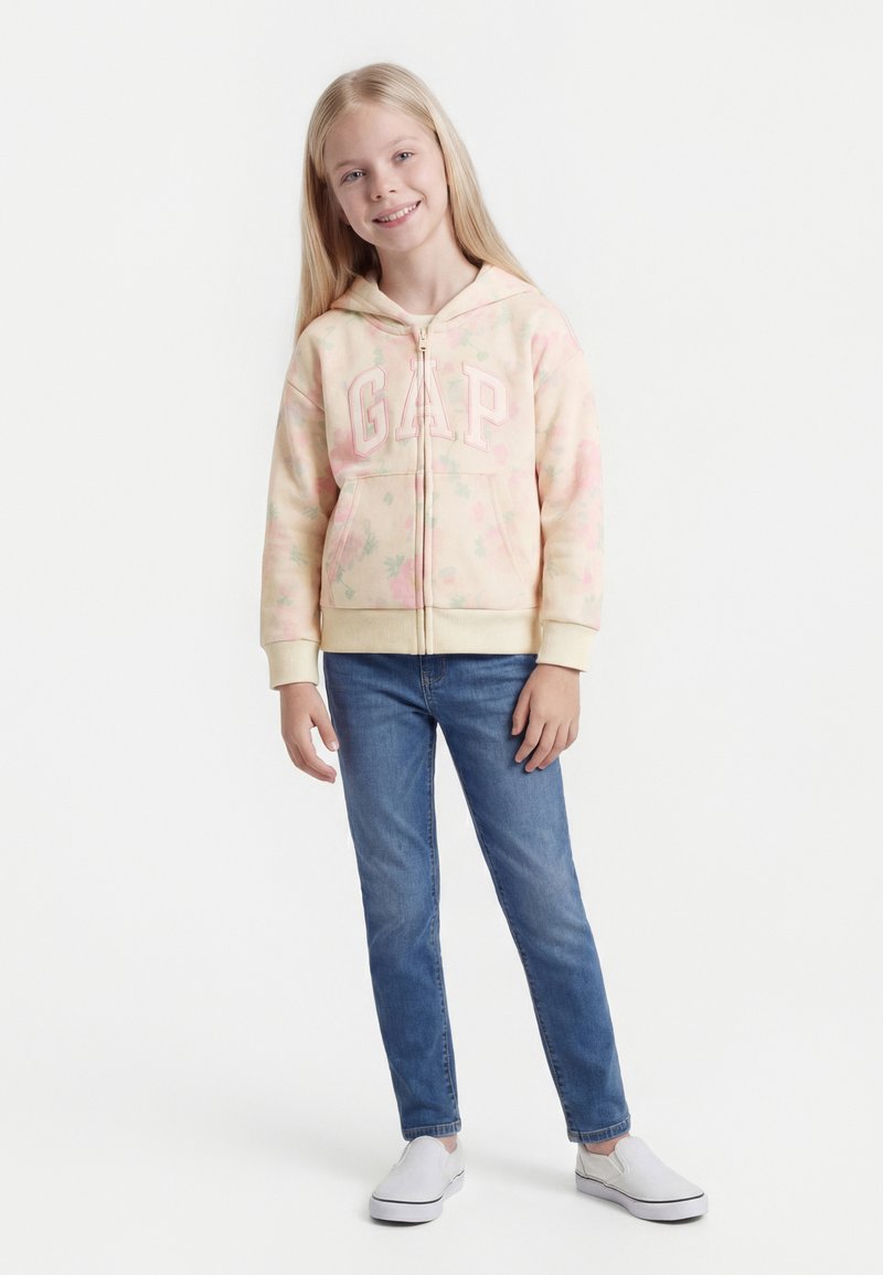 Jeune fille aux longs cheveux blonds portant un sweat à capuche GAP tie-dye pastel, un jean bleu et des chaussures blanches à enfiler, debout et souriante.