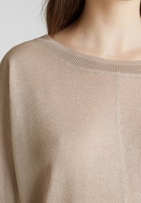 Beżowy sweter z dzianiny z okrągłym dekoltem noszony przez osobę z długimi brązowymi włosami, odsłaniający górną część klatki piersiowej i ramienia.