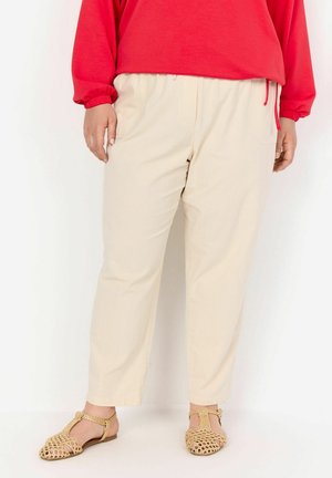 Cremfarbene, lässig geschnittene Hose mit einem elastischen Bund und Taschen, kombiniert mit beigen, gewebten Sandalen und einem roten Langarm-Oberteil.