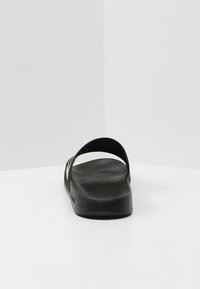 Svart gummistövla med två tunna remmar och en strukturerad fotbädd. Klackhöjden är måttlig och har en rundad tådesign.