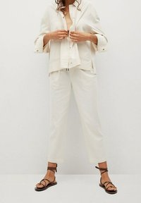 Lichtbeige linnen outfit met een losse jas met knopen en elastische taillebroek, aangevuld met bruine sandalen met banden.