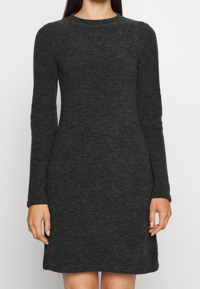 Femme portant une robe en maille gris foncé à manches longues, longueur au genou, debout devant un fond clair uni.