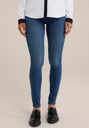 Jeans ajustados en denim azul oscuro con una textura desgastada. Combinados con mocasines negros. El conjunto incluye una blusa blanca con detalles en negro.
