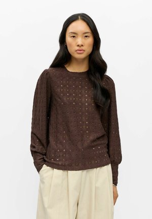 Femme aux longs cheveux foncés portant un chemisier marron texturé à manches longues et un pantalon beige ample, debout devant un fond blanc.