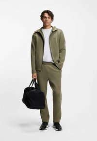 Oliwkowy sportowy dres z kurtką na suwak i spodniami, w połączeniu z czarną torbą podróżną i czarnymi butami do biegania. Prosty design, bez wzorów.