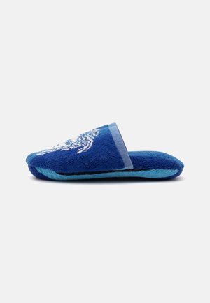 Blå slip-on tøfler med blødt stof og et hvidt mønster på toppen, vist fra siden på en ensfarvet baggrund.