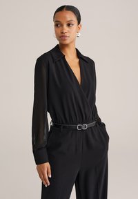 Čierny overal s prekrývací konštrukciou, priehľadnými rukávmi a kontrastným opaskom. Má golier a bočné vrecká. Hladká textura.