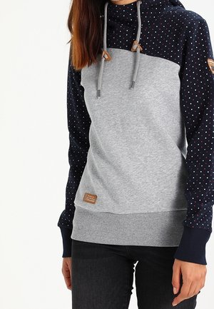 Kobieta ubrana w szarą i granatową bluzę z kapturem z małymi kropkami na rękawach i górnej części klatki piersiowej, w połączeniu z czarnymi spodniami.