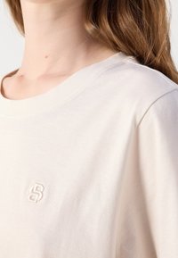 Gros plan d'une personne portant une chemise blanc cassé avec un logo "SB" cousu sur la poitrine et de longs cheveux châtain clair tombant sur l'épaule.