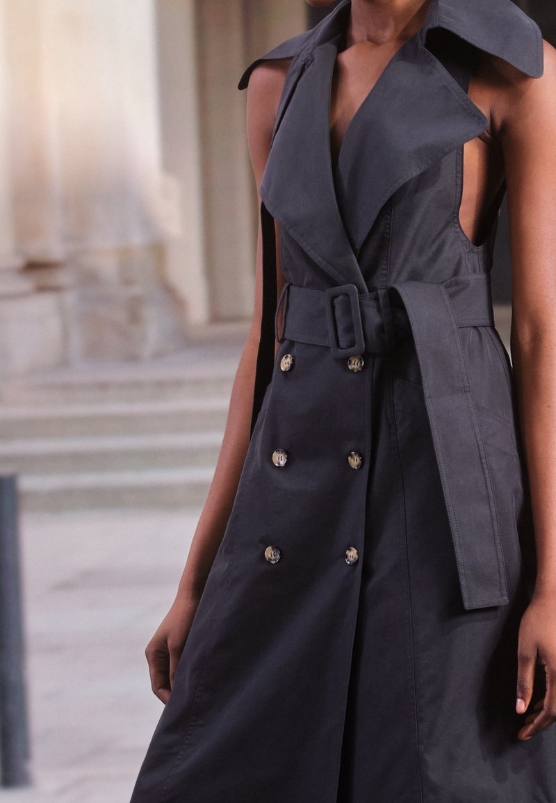 Trench-coat noir avec taille ceinturée, boutons croisés, large col et découpes latérales, confectionné dans un tissu lisse à finition mate.
