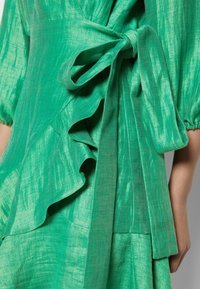 Robe verte en tissu texturé avec manches bouffantes, un grand nœud noué à la taille et des détails froncés sur le côté.