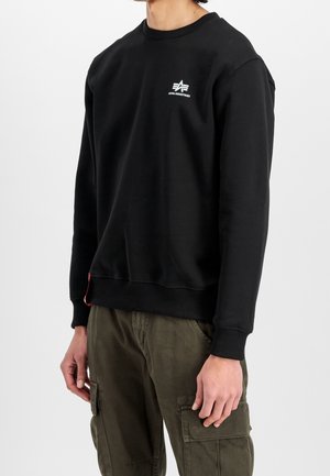 Homme portant un sweat-shirt noir à col ras du cou avec un petit logo blanc Alpha Industries sur la poitrine et un pantalon cargo vert olive.