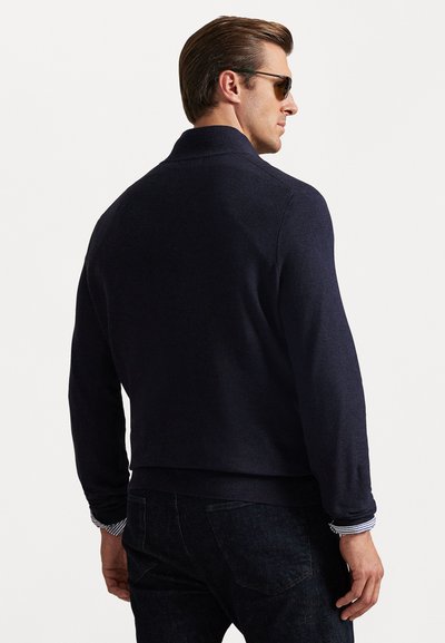 Σκούρα μπλούζα με γιακά, υφασμένη και με μακριά μανίκια. Περιλαμβάνει ριγέ λεπτομέρεια στον λαιμό του μανικιού. Φοράει με σκούρα τζιν.