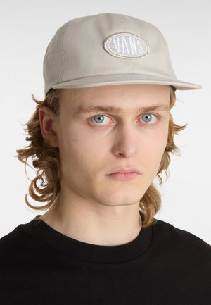 Beige pet gemaakt van katoen, met een witte geborduurde logopatch op de voorkant, een vlakke rand en een gestructureerd oppervlak.