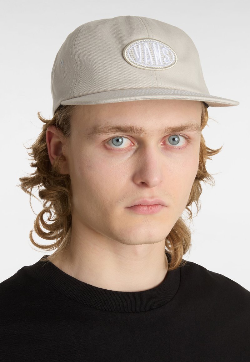 Beige cap made of cotton, featuring a white embroidered logo patch on the front, with a flat brim and textured surface.