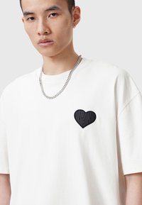 Camiseta de algodón blanca con un parche negro texturizado en forma de corazón en el lado izquierdo del pecho, mangas cortas y un corte relajado, combinada con una cadena de plata.