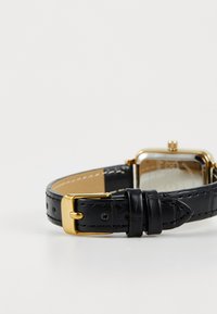 Reloj con correa de cuero negro y hebilla dorada. La correa presenta detalles de costura y agujeros. La caja es cuadrada con un acabado en tono dorado.