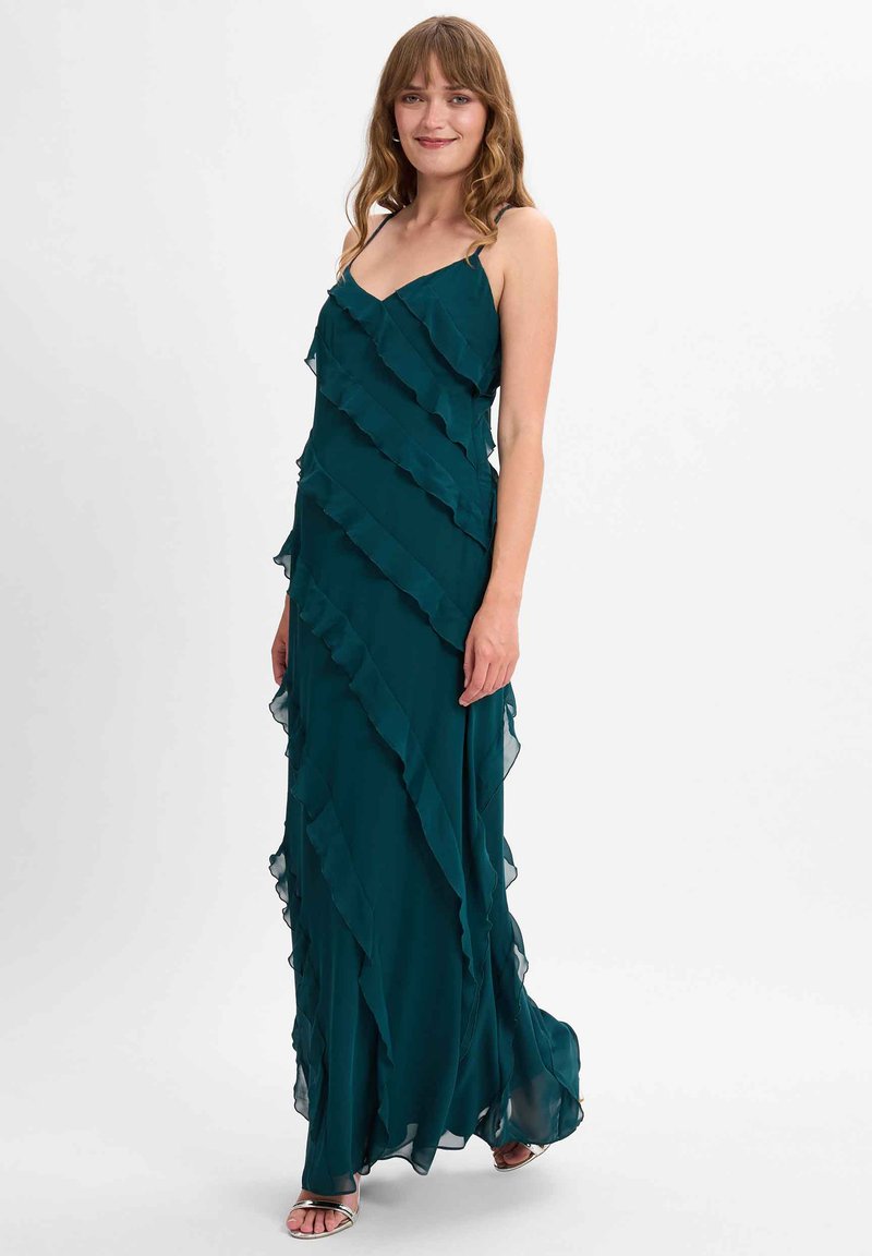 Tiefblaue Maxi-Kleid mit geschichtetem Rüschendesign, verstellbaren Spaghetti-Trägern, fließendem Stoff und kniehohem Unterrock, kombiniert mit silbernen Sandalen.