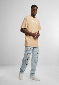 Beige cotton T-shirt with short sleeves featuring white text. Light blue ripped jeans with multiple distressed patches. White sneakers.
