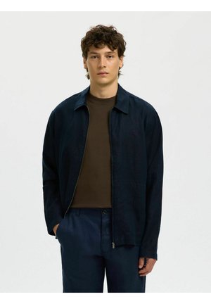 Giovane uomo con capelli corti e ricci che indossa una giacca con cerniera blu scuro sopra una maglietta marrone e pantaloni blu scuro abbinati, in piedi contro uno sfondo neutro.