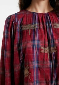 Blouse à carreaux rouges ornée d'une broderie dorée complexe de libellules et d'un élément décoratif en bas à gauche. Encolure et manches froncées.