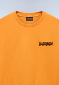 Oranssi puuvillapaita, jossa on pyöreä pääntie ja pieni musta "NAPAPIJRI geographic" -logo vasemmassa rinnassa. Liukas pinta.