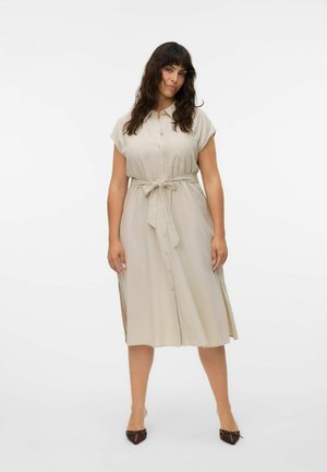 Kvinde med langt mørkt hår iført en beige midikjole med knapper, korte ærmer, sidelommer og et bindebælte i taljen, kombineret med mørke spidse flade sko.