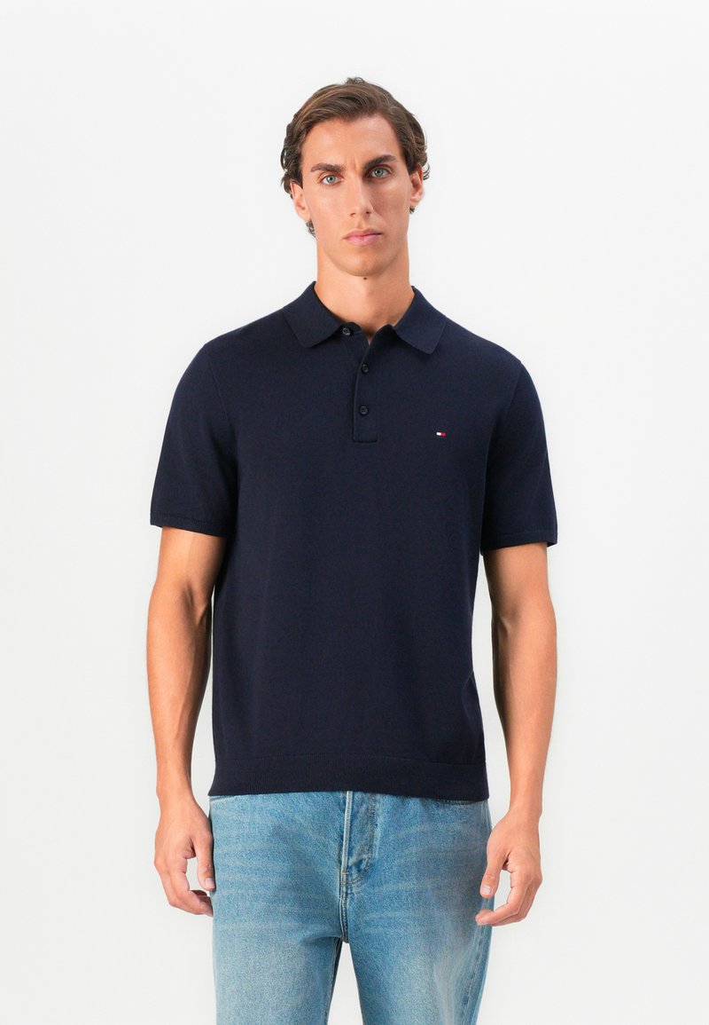 Mladý muž v tmavomodrej polokošeli s krátkym rukávom a gombíkmi, v svetlomodrých džínsoch, stojaci pred jednoliatym bielym pozadím.