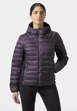 Mujer con chaqueta acolchada con capucha de color púrpura oscuro con cremallera frontal, de pie con una mano en el bolsillo y expresión neutral.