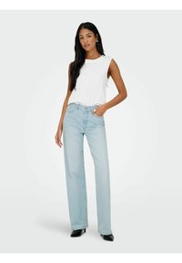 Λευκή αμάνικη μπλούζα συνδυασμένη με ελαφρώς ξεθωριασμένα, ψηλόμεσα, φαρδιά παντελόνια. Μοντέλο που φοράει μαύρα ψηλοτάκουνα παπούτσια, παρουσιάζοντας μια casual ενδυμασία.