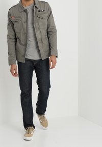 Grå, let jakke med knaplukning og skulderlapper, båret over en lysegrå skjorte; parret med mørkeblå jeans og beige sneakers.