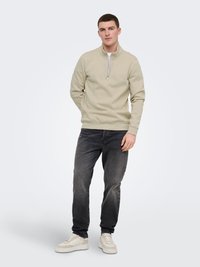 Jeune homme debout les mains dans les poches, portant un sweat-shirt beige à fermeture éclair quart, un jean noir délavé et des baskets blanches sur fond uni.